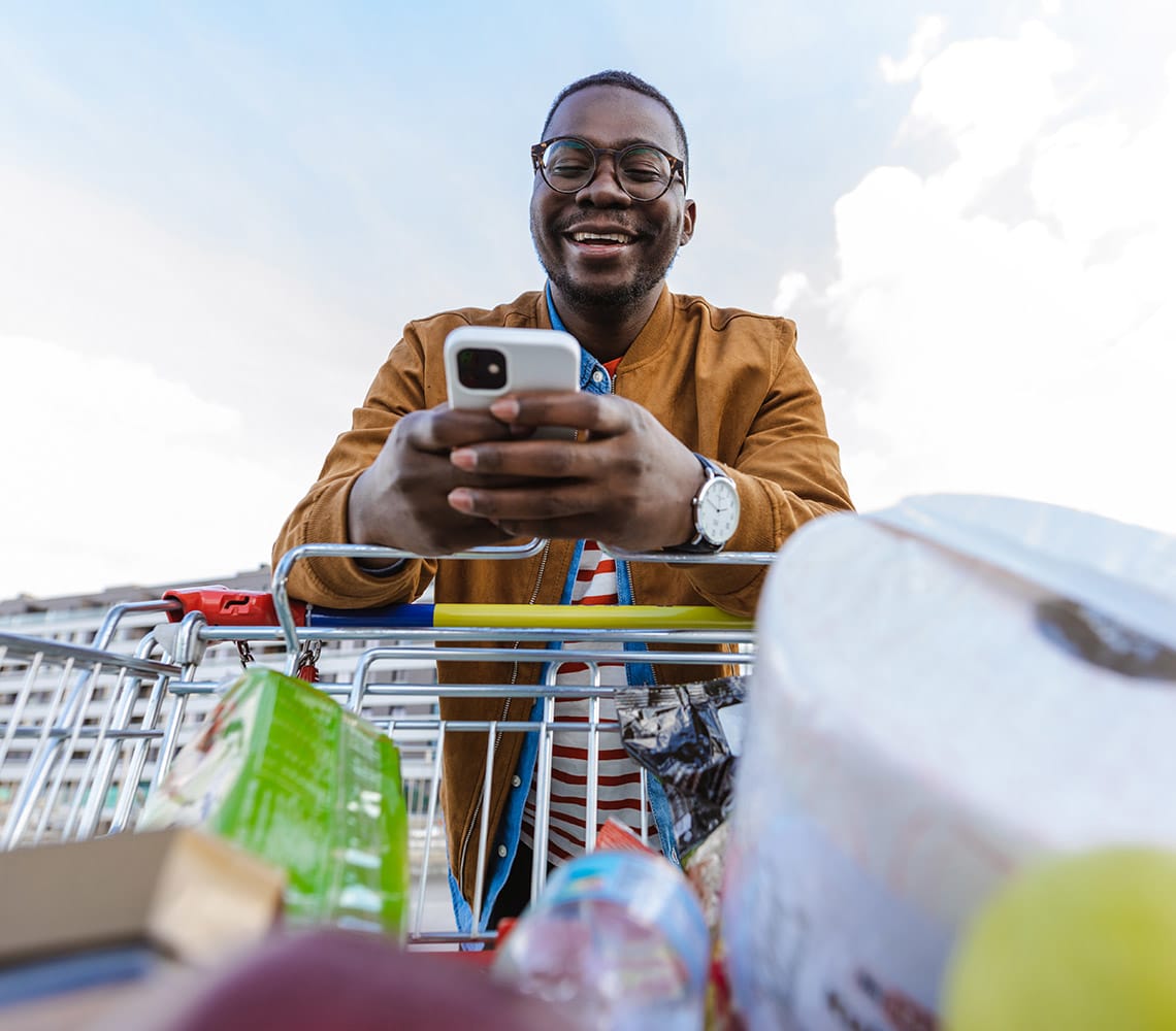 A person in a brown jacket smiles and looks at their smartphone while leaning on a shopping cart filled with groceries.